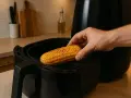En hand placerar en gyllene mazorca de maiz i en airfryer.