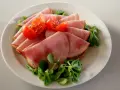 Skivor av saftig skinka, en köttprodukt, serveras med körsbärstomater och gröna blad på en tallrik.
