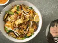 En skål med nudlar, tofu, svamp och grönsaker. En kvinna ler i ett hörn.