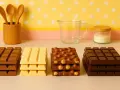 Fyra staplar med olika tipos de chocolates: mörk, vit, med nötter och mörk igen. En träbehållare med skedar, ett glas och en burk med socker i bakgrunden.