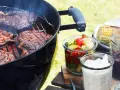 Grillad pernil de cerdo ryker på grillen. Majskolvar, sallad och såser står redo för en härlig sommarmåltid.