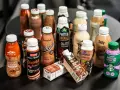 En samling flaskor och kartonger med olika sorters batido de proteinas, mestadels chokladsmak.