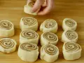 Färska kanelbullar, eller "rollo de canela", ligger redo att bakas på en träskärbräda. En hand placerar en bullform.