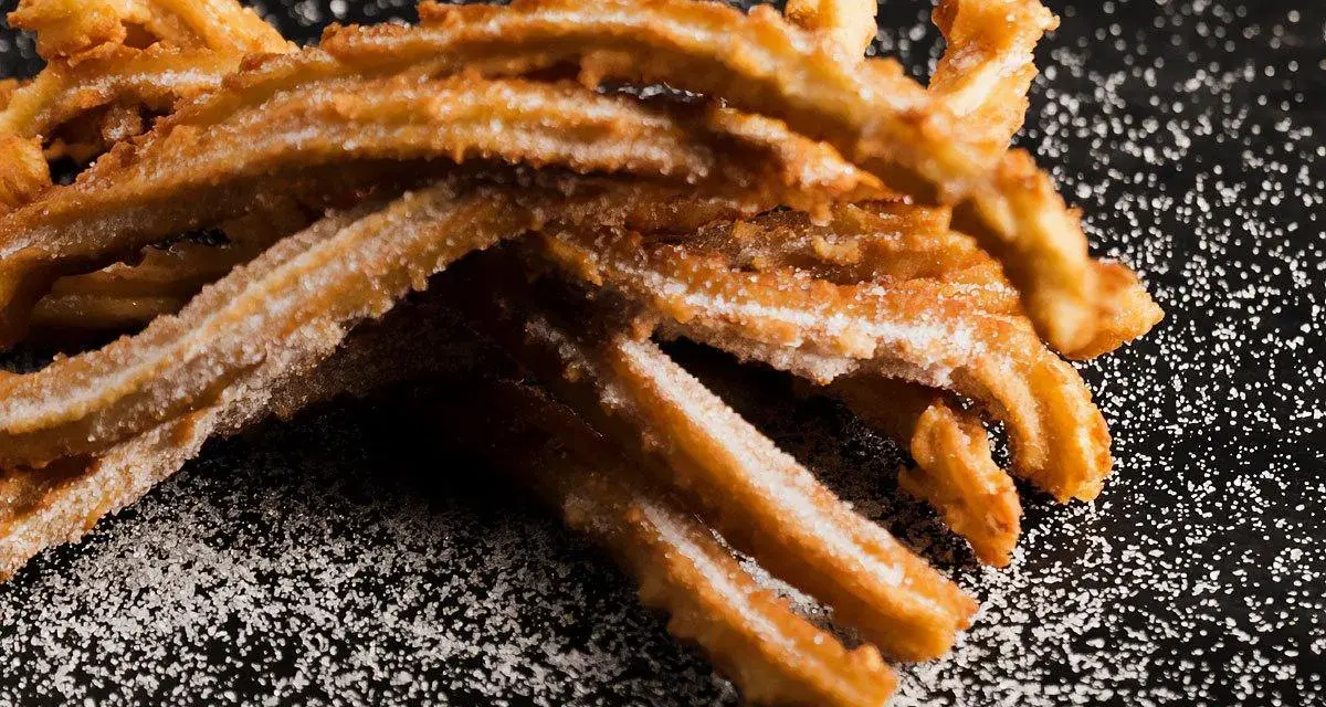 En hög med gyllene churros españoles, pudrade med socker, ligger på en mörk yta.
