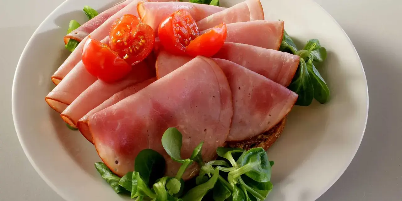 Skivor av saftig skinka, en köttprodukt, serveras med körsbärstomater och gröna blad på en tallrik.