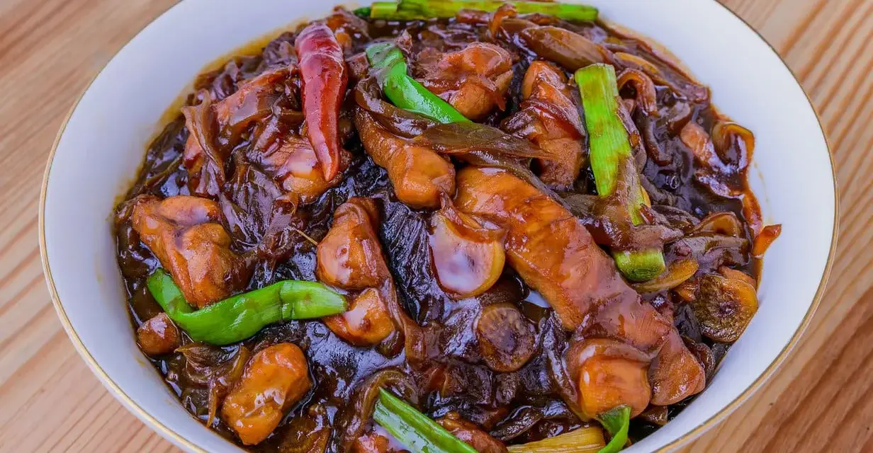 En skål med läcker pollo encebollado, med saftiga kycklingbitar i en mörk sås, toppad med gröna salladslökar och en röd chili.