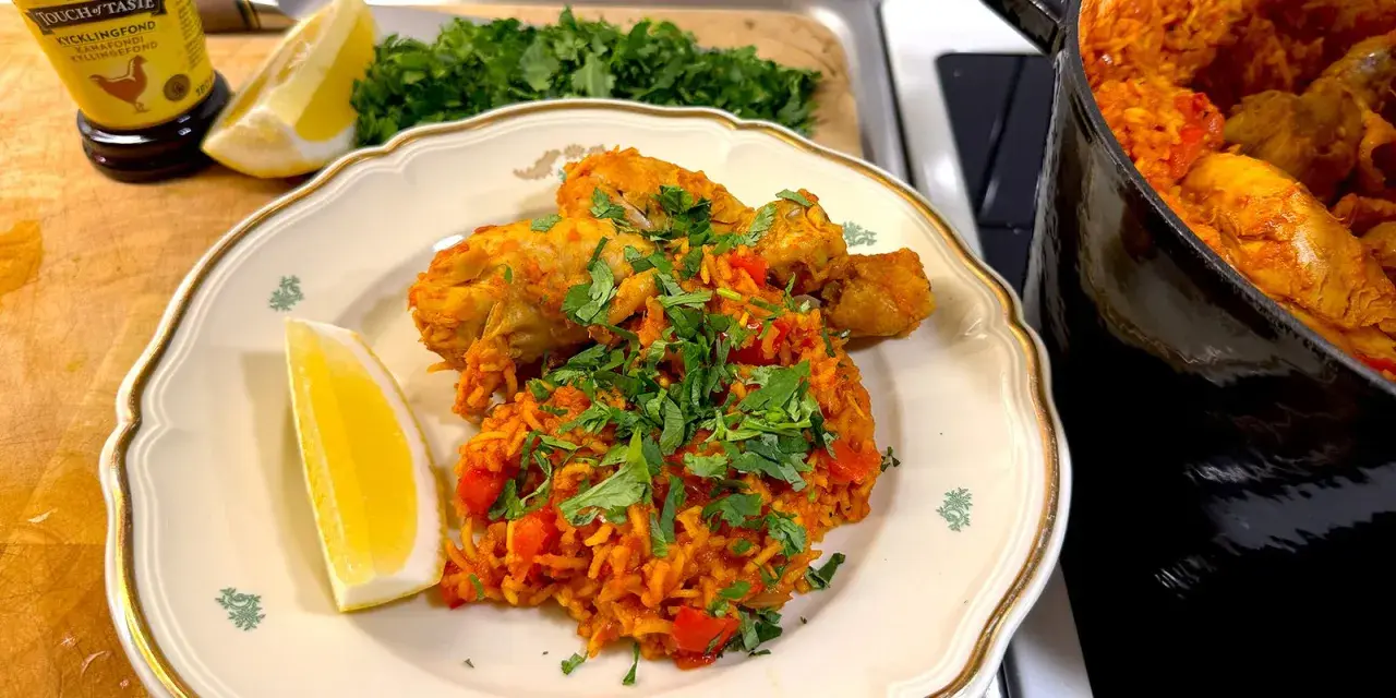 En tallrik med kyckling och ris, kryddad med paprika och persilja. En citronklyfta ligger bredvid. En gryta med samma rätt syns i bakgrunden.