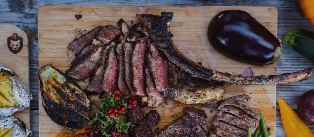 En saftig tomahawk steak, skuren i skivor, serveras med grillad aubergine, potatis i folie och lingon.