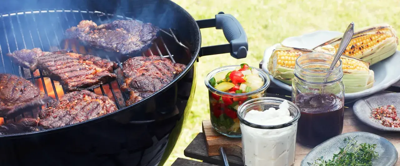 Grillad pernil de cerdo ryker på grillen. Majskolvar, sallad och såser står redo för en härlig sommarmåltid.