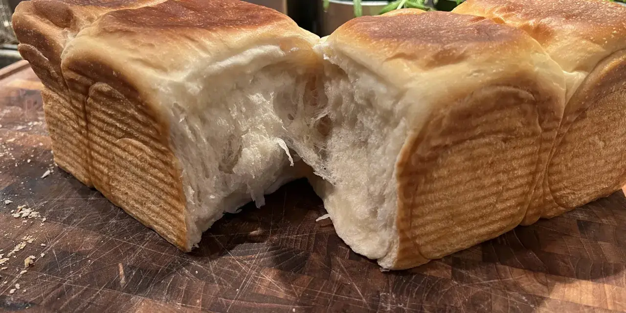 Mjukt och fluffigt **pan de leche** som brutits isär på en träskärbräda. Den gyllene skorpan kontrasterar mot det vita, trådiga inkråmet.