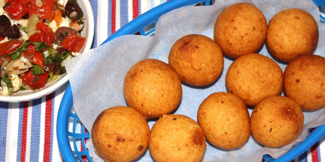 Gylla gyllenbruna friterade bollar, som visar hur man gör buñuelos, i en blå korg. Bredvid en skål med en färgglad sallad.