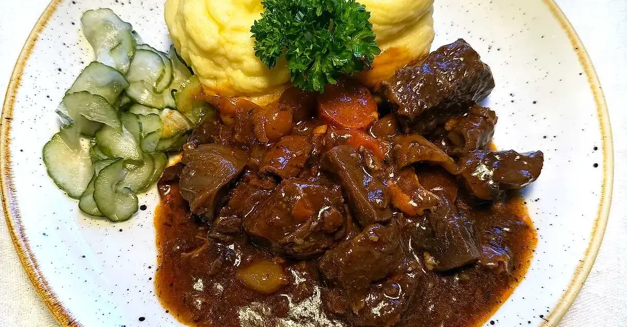 En tallrik med mustigt estofado de res, krämigt potatismos och skivad gurka. En grön persiljekvist toppar moset.