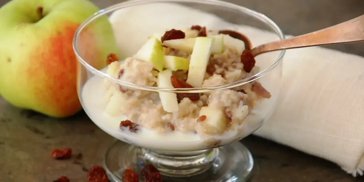En skål med havregrynsgröt med äppelbitar och russin, bredvid ett äpple. Enkel guide för como preparar avena.