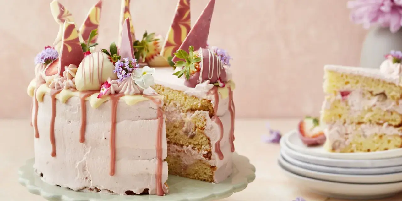 En rosa tårta med jordgubbsfrosting, chokladdekorationer och färska bär. En bit av tårtan visas på en stapel tallrikar.