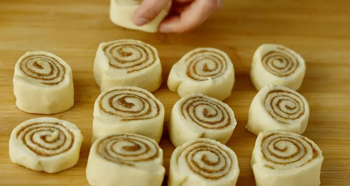 Färska kanelbullar, eller "rollo de canela", ligger redo att bakas på en träskärbräda. En hand placerar en bullform.