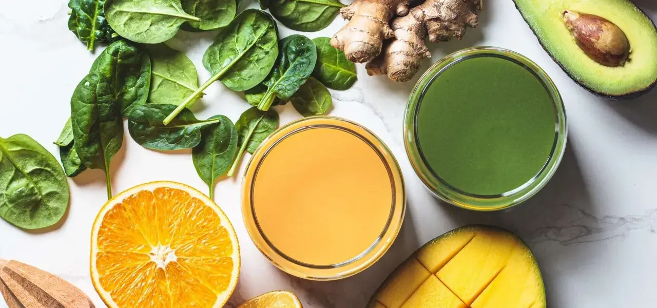 Färgglada juicer, en med apelsin och en grön, omgivna av färska ingredienser som spenat, ingefära, mango och avokado. Perfekt för en hälsosam "te de curcuma y jengibre"-inspirerad smoothie.