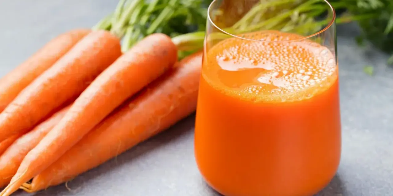 Färskt morotsjuice i ett glas, bredvid en hög med morötter. Morotsjuice är bra för synen och immunförsvaret.