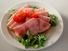 Skivor av saftig skinka, en köttprodukt, serveras med körsbärstomater och gröna blad på en tallrik.