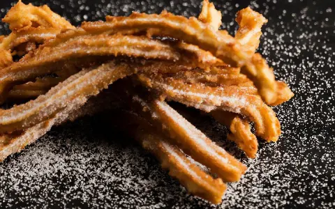 En hög med gyllene churros españoles, pudrade med socker, ligger på en mörk yta.