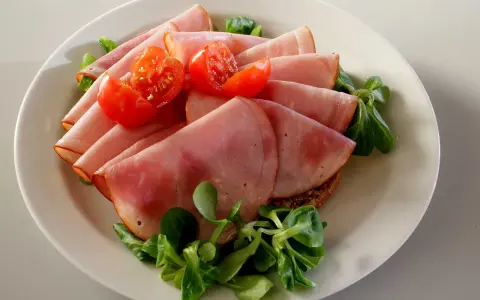 Skivor av saftig skinka, en köttprodukt, serveras med körsbärstomater och gröna blad på en tallrik.