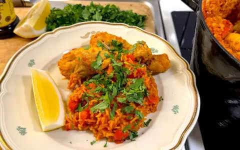 En tallrik med kyckling och ris, kryddad med paprika och persilja. En citronklyfta ligger bredvid. En gryta med samma rätt syns i bakgrunden.