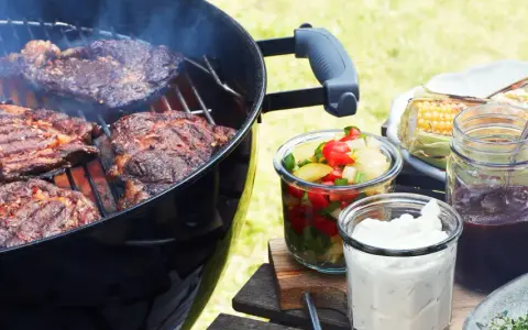 Grillad pernil de cerdo ryker på grillen. Majskolvar, sallad och såser står redo för en härlig sommarmåltid.