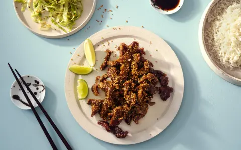 Krispiga chicharrones de cerdo med sesamfrön, serverade med ris, sallad och limeklyftor.