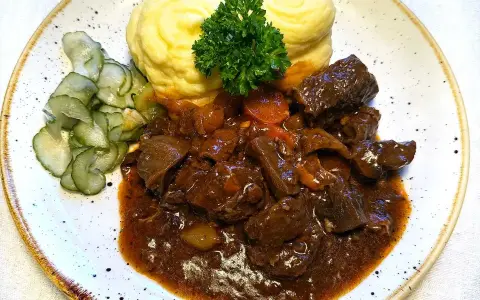 En tallrik med mustigt estofado de res, krämigt potatismos och skivad gurka. En grön persiljekvist toppar moset.