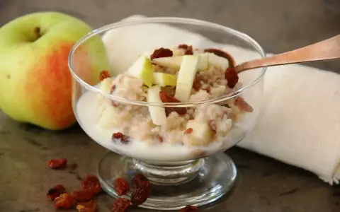 En skål med havregrynsgröt med äppelbitar och russin, bredvid ett äpple. Enkel guide för como preparar avena.