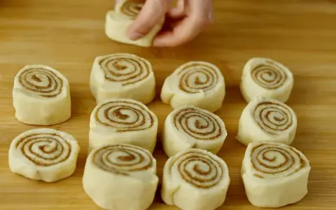 Färska kanelbullar, eller "rollo de canela", ligger redo att bakas på en träskärbräda. En hand placerar en bullform.