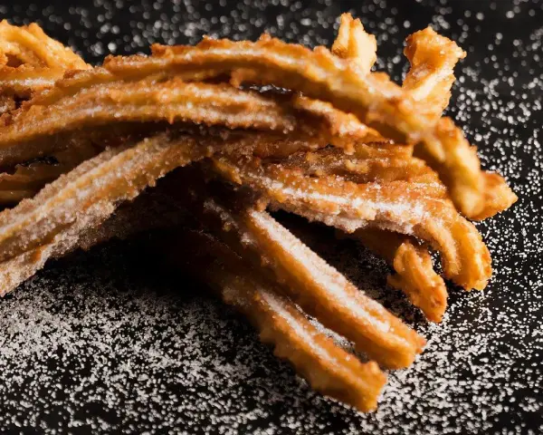 En hög med gyllene churros españoles, pudrade med socker, ligger på en mörk yta.