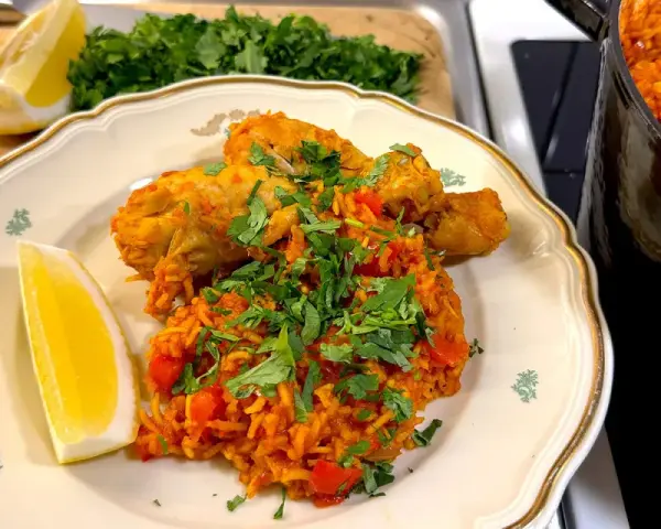 En tallrik med kyckling och ris, kryddad med paprika och persilja. En citronklyfta ligger bredvid. En gryta med samma rätt syns i bakgrunden.