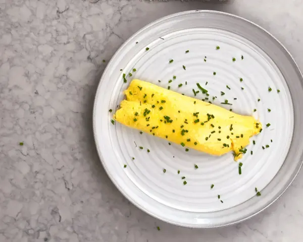 En perfekt rullad omelett, strödd med gräslök, på en tallrik. Enkel guide för como hacer un omelet.