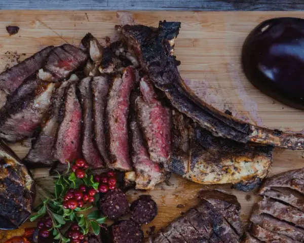 En saftig tomahawk steak, skuren i skivor, serveras med grillad aubergine, potatis i folie och lingon.