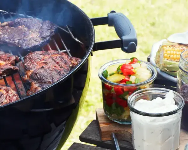 Grillad pernil de cerdo ryker på grillen. Majskolvar, sallad och såser står redo för en härlig sommarmåltid.