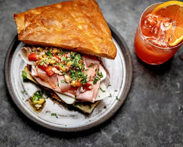 En saftig macka med pan integral, skinka och grönsaker, serverad med en orange drink med apelsinskiva.