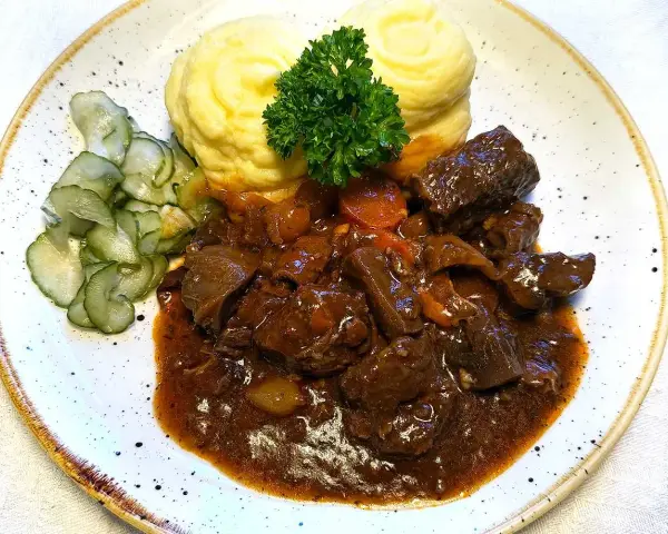 En tallrik med mustigt estofado de res, krämigt potatismos och skivad gurka. En grön persiljekvist toppar moset.