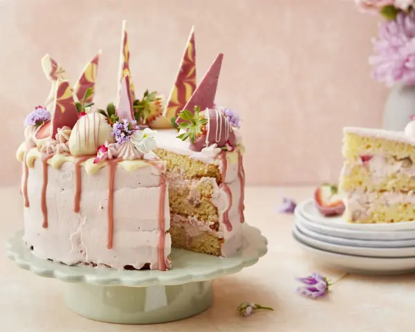 En rosa tårta med jordgubbsfrosting, chokladdekorationer och färska bär. En bit av tårtan visas på en stapel tallrikar.