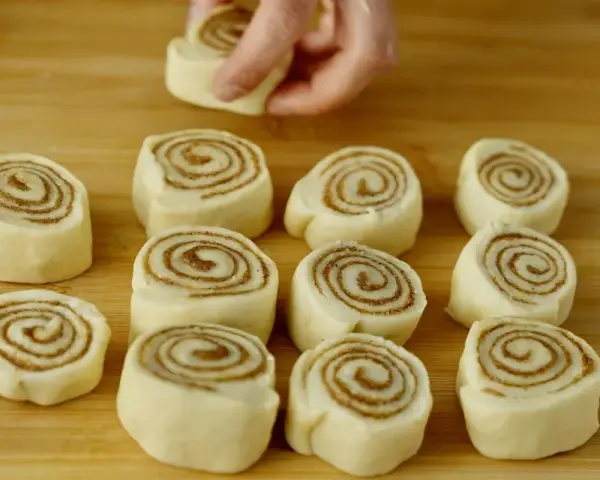 Färska kanelbullar, eller "rollo de canela", ligger redo att bakas på en träskärbräda. En hand placerar en bullform.