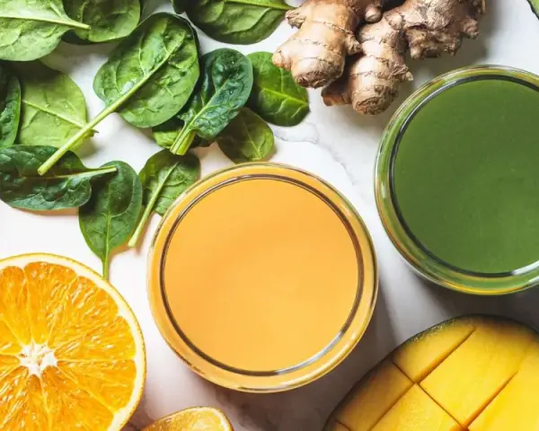 Färgglada juicer, en med apelsin och en grön, omgivna av färska ingredienser som spenat, ingefära, mango och avokado. Perfekt för en hälsosam "te de curcuma y jengibre"-inspirerad smoothie.