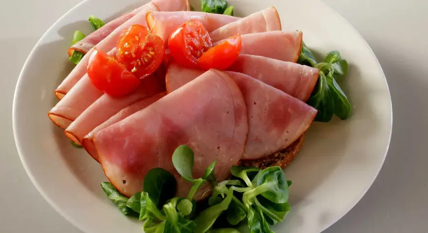 Skivor av saftig skinka, en köttprodukt, serveras med körsbärstomater och gröna blad på en tallrik.
