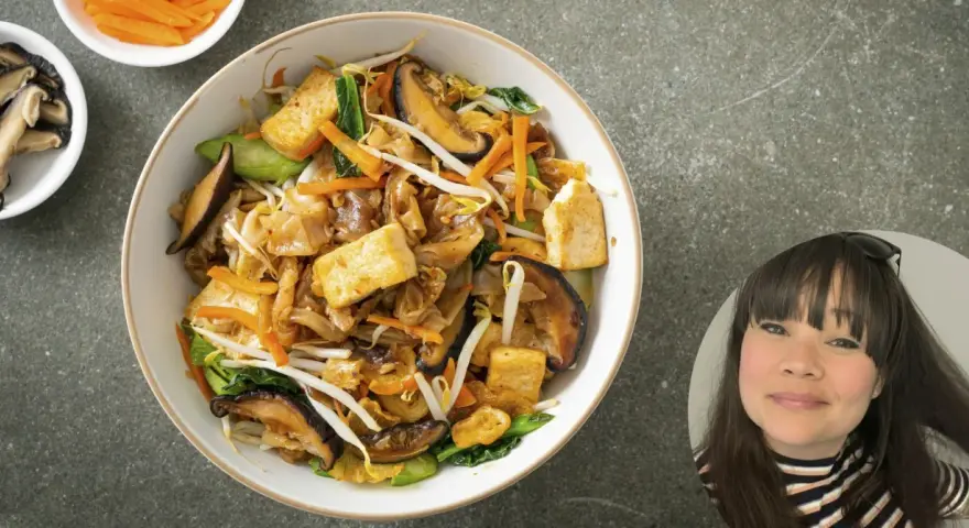 En skål med nudlar, tofu, svamp och grönsaker. En kvinna ler i ett hörn.