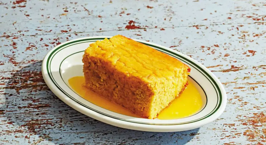 En bit gyllene pan de maiz, saftig och serverad med en gyllene sirap på en vit tallrik med gröna kanter.