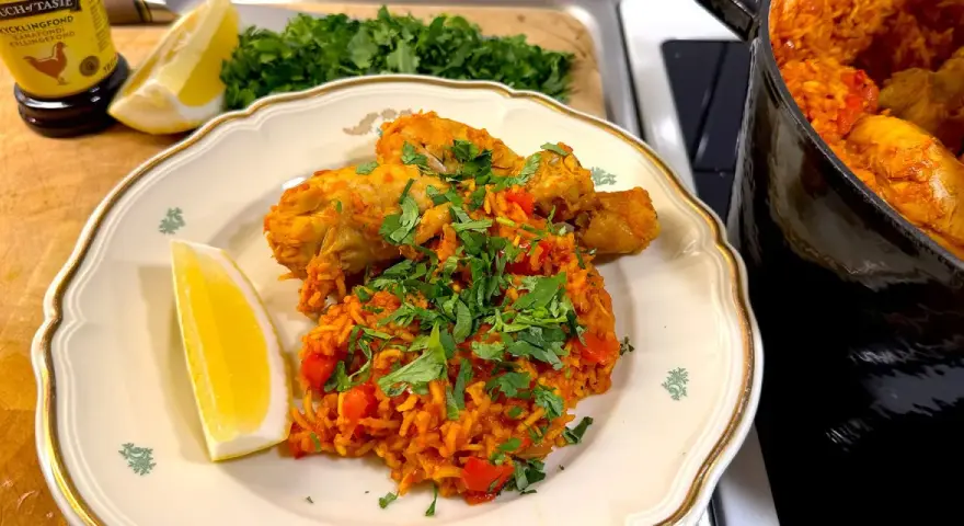 En tallrik med kyckling och ris, kryddad med paprika och persilja. En citronklyfta ligger bredvid. En gryta med samma rätt syns i bakgrunden.