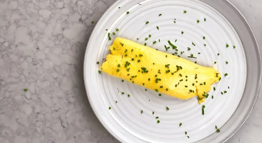En perfekt rullad omelett, strödd med gräslök, på en tallrik. Enkel guide för como hacer un omelet.