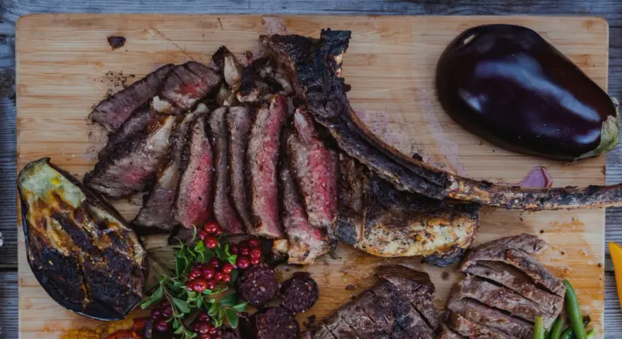 En saftig tomahawk steak, skuren i skivor, serveras med grillad aubergine, potatis i folie och lingon.