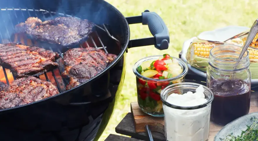 Grillad pernil de cerdo ryker på grillen. Majskolvar, sallad och såser står redo för en härlig sommarmåltid.