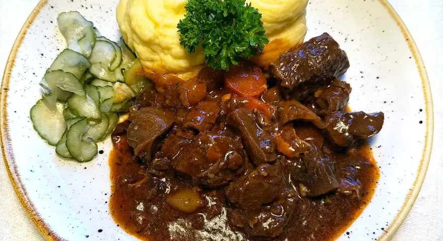 En tallrik med mustigt estofado de res, krämigt potatismos och skivad gurka. En grön persiljekvist toppar moset.