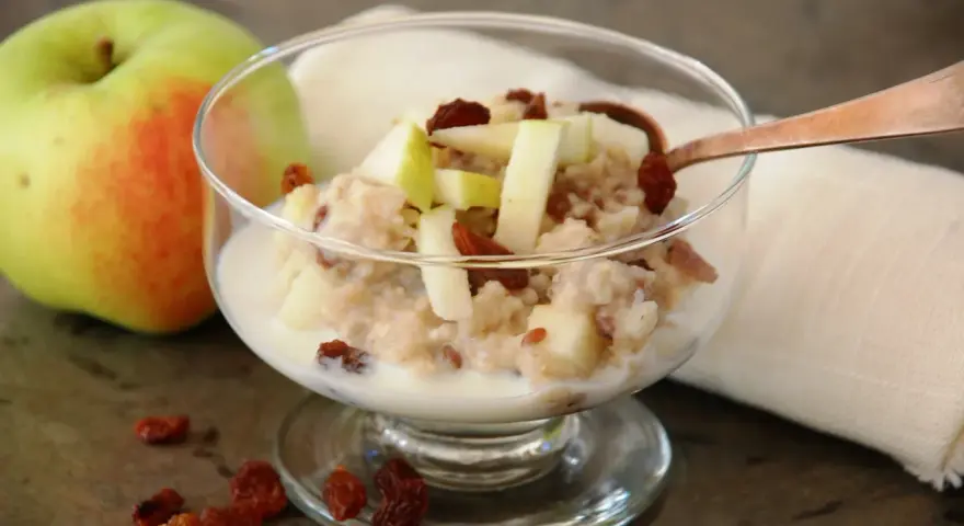 En skål med havregrynsgröt med äppelbitar och russin, bredvid ett äpple. Enkel guide för como preparar avena.