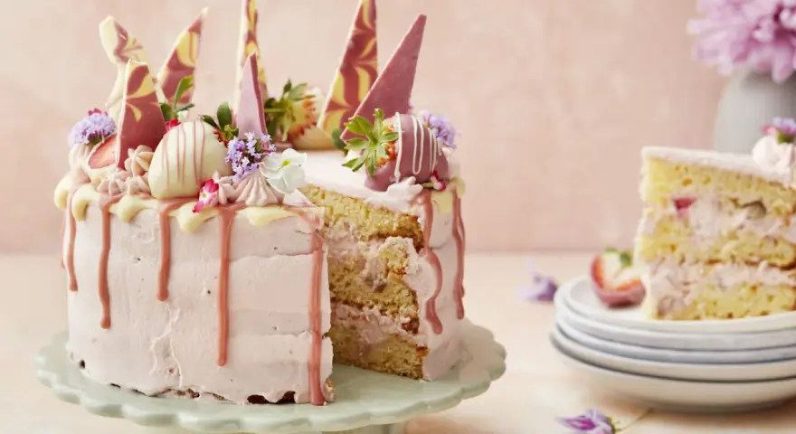 En rosa tårta med jordgubbsfrosting, chokladdekorationer och färska bär. En bit av tårtan visas på en stapel tallrikar.